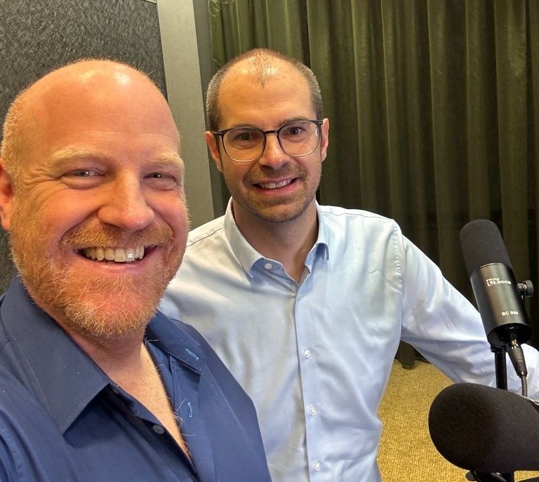 Richard Seidl und Matthias Groß lächeln gemeinsam in die Kamera bei der Aufnahme der Podcast-Folge, im Vordergrund ein Studiomikrofon.