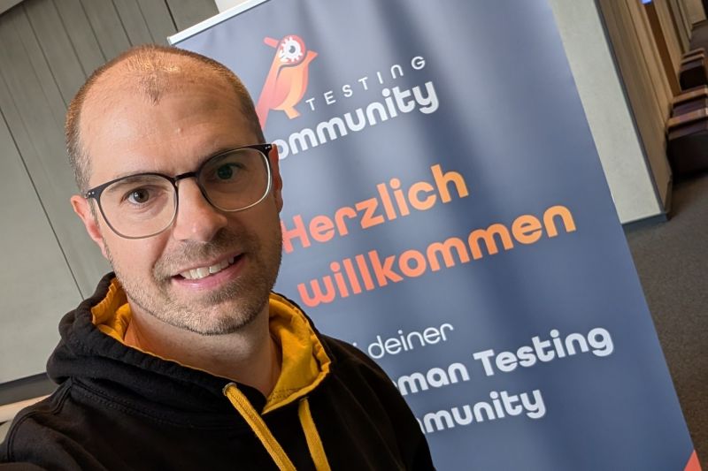 Selfie von Matthias Groß im schwarzen TestLänd-Kapuzenpulli vor dem Roll-up der German Testing Community mit der Aufschrift Herzlich willkommen, beim German Testing Day 2026 im Kap Europa in Frankfurt.