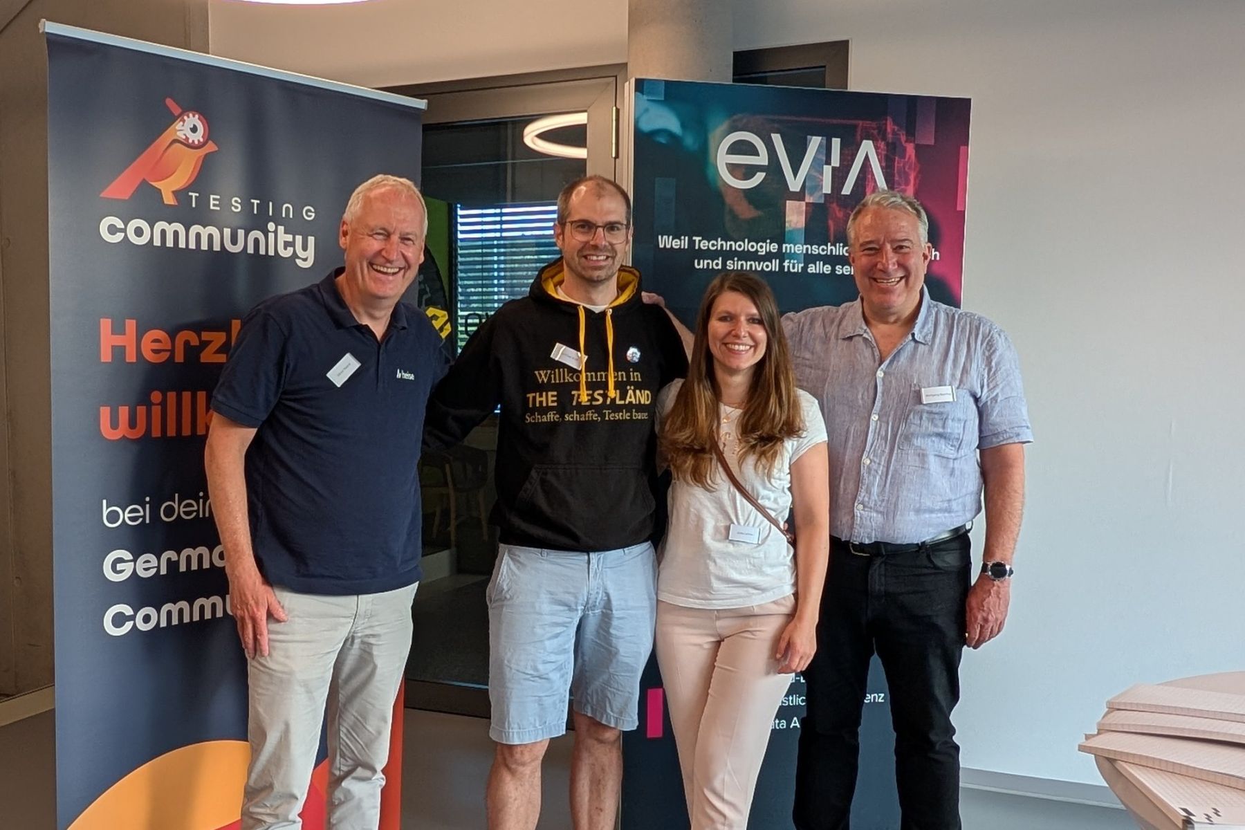 Gruppenbild bei der German Testing Night in Leonberg: Matthias Groß im schwarzen TestLänd-Kapuzenpulli mit 3 weiteren lachenden Personen vor den Roll-ups der German Testing Community und von evia.