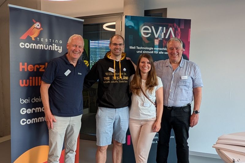 Gruppenbild bei der German Testing Night in Leonberg: Matthias Groß im schwarzen TestLänd-Kapuzenpulli mit 3 weiteren lachenden Personen vor den Roll-ups der German Testing Community und von evia.