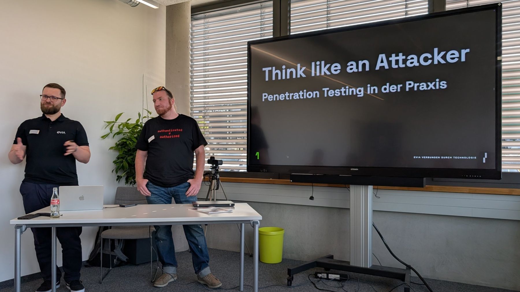 2 Vortragende stehen neben einem Tisch mit Laptop vor einem großen Bildschirm mit der Titelfolie Think like an Attacker: Penetration Testing in der Praxis.