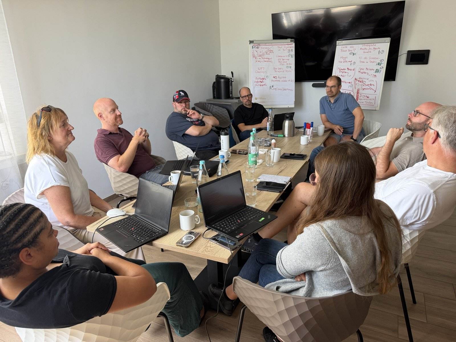 Diskussionsrunde am Tisch mit Laptops, Kaffeetassen und Wasserflaschen, im Hintergrund 2 Flipcharts mit den gesammelten Themen und Strichlisten zur Priorisierung.