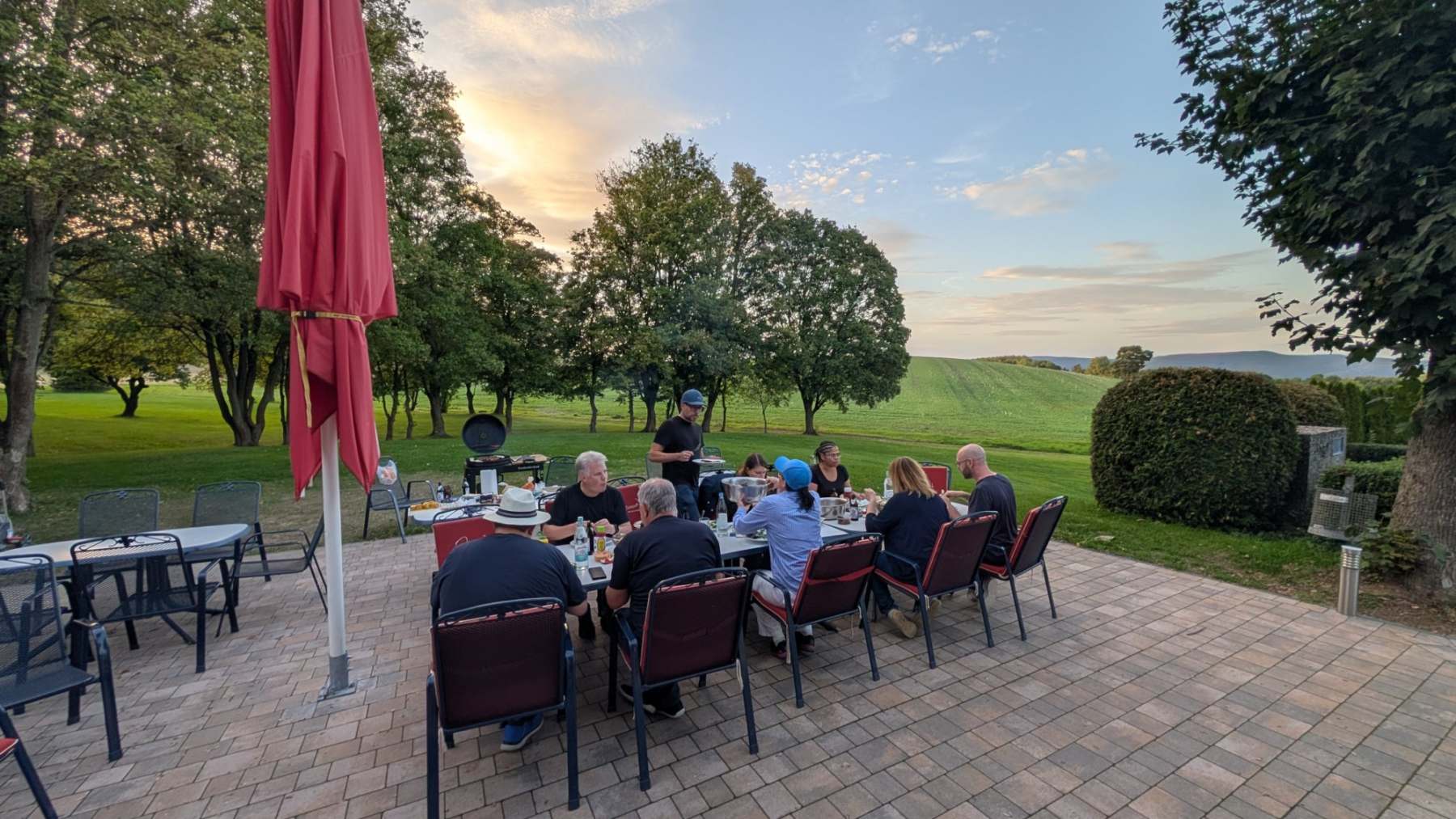 Teilnehmende essen an einer langen Tafel auf der Terrasse, eine Person bringt Gegrilltes vom Grill, im Hintergrund Bäume, Felder und der Sonnenuntergang.