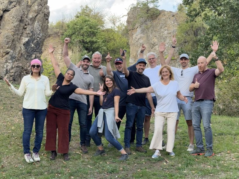 Gruppenbild beim 2. German Testing Retreat: gut gelaunte Teilnehmende mit erhobenen Armen auf einer Wiese vor Felswänden an der Blauen Kuppe bei Eschwege.