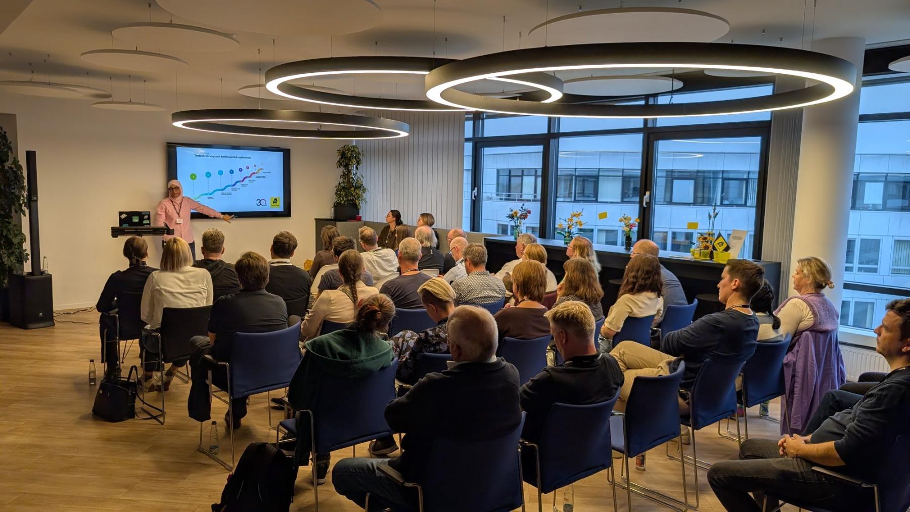 Lilia Gargouri zeigt beim TestLänd After Work #6 eine Folie zur Optimierung der Testausführungszeit vor voll besetzten Stuhlreihen.