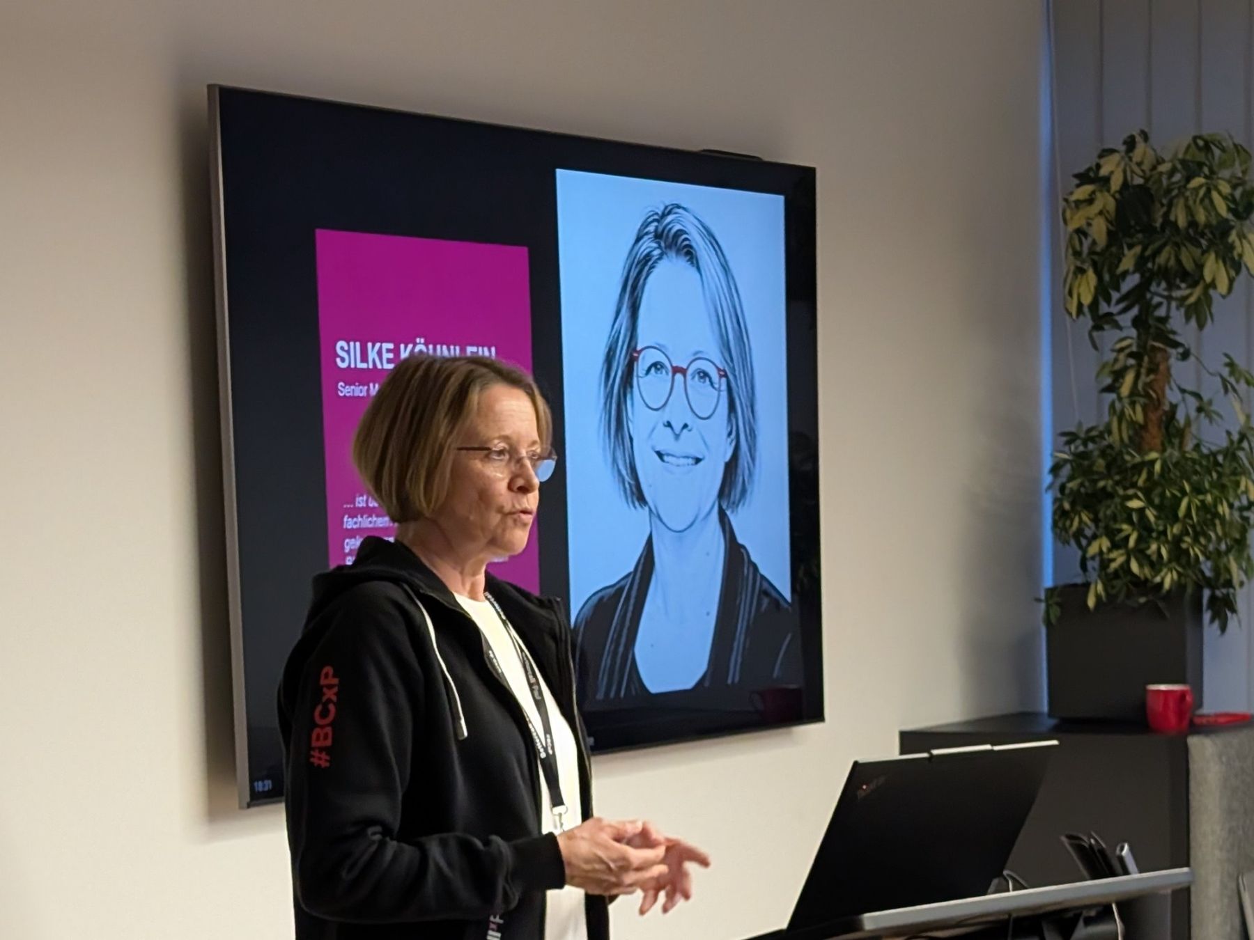 Silke Köhnlein hält beim TestLänd After Work #6 ihren Vortrag über Fachwissen, Anforderungen und KI im Test, hinter ihr auf dem Bildschirm ihre Vorstellungsfolie mit einer Porträtzeichnung.