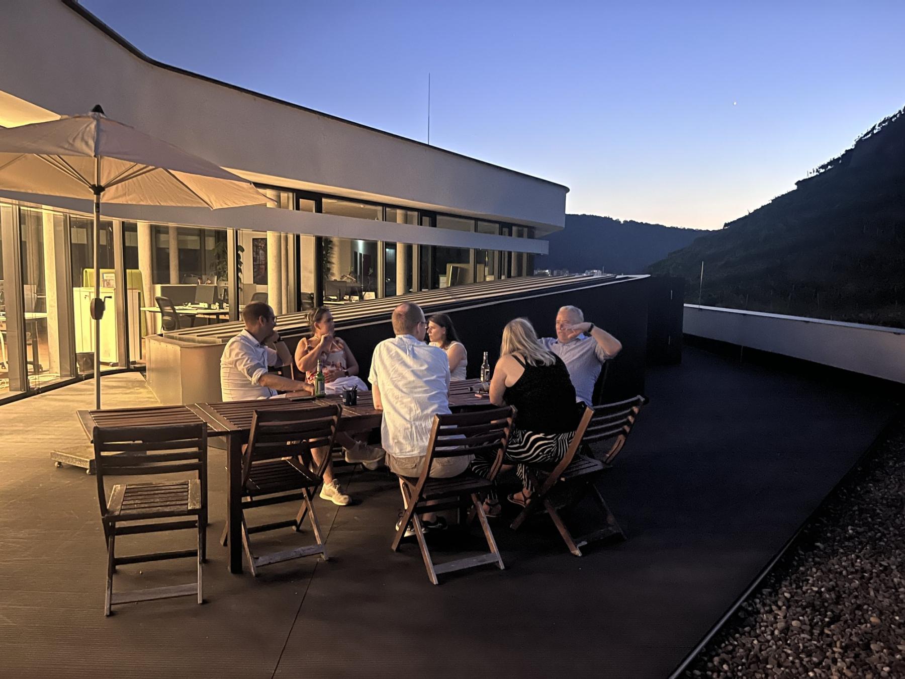 Eine Runde Teilnehmender sitzt in der Dämmerung am Tisch auf der Dachterrasse und diskutiert, dahinter das beleuchtete Bürogebäude.