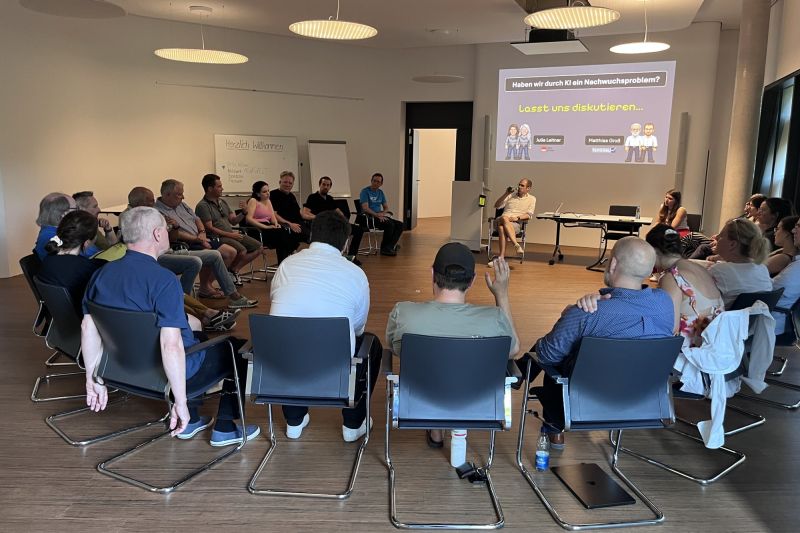 Teilnehmende des TestLänd After Work im Stuhlkreis, eine Person meldet sich zu Wort, auf der Leinwand die Frage: Haben wir durch KI ein Nachwuchsproblem?