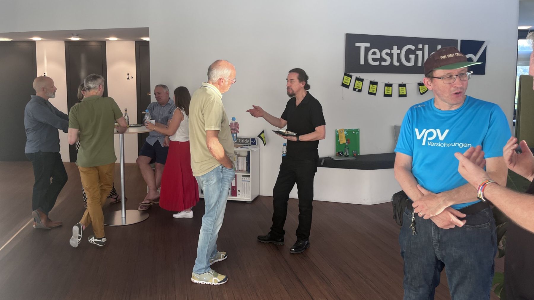 Teilnehmende im Gespräch im Foyer der TestGilde, an der Wand das TestGilde-Logo und eine Wimpelkette vom TestLänd.