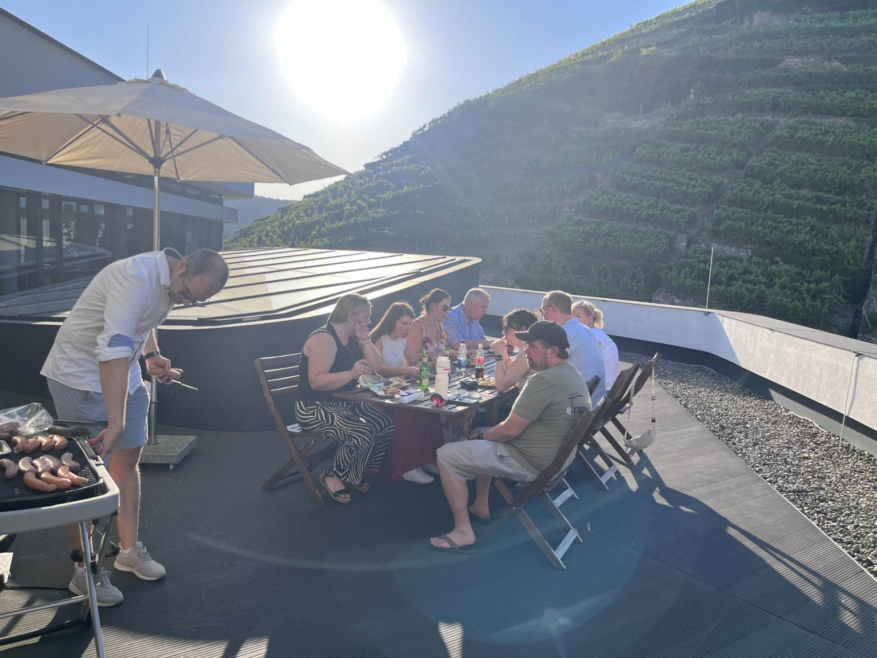 Eine Person grillt Würste, daneben essen Teilnehmende an einem Holztisch auf der Dachterrasse der TestGilde in Esslingen, im Hintergrund die Weinberge in der Abendsonne.