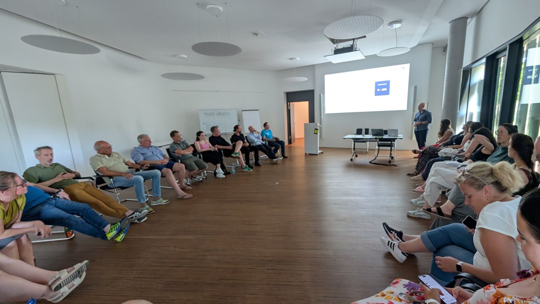Teilnehmende sitzen im Stuhlkreis, ein Vortragender steht neben der Leinwand.