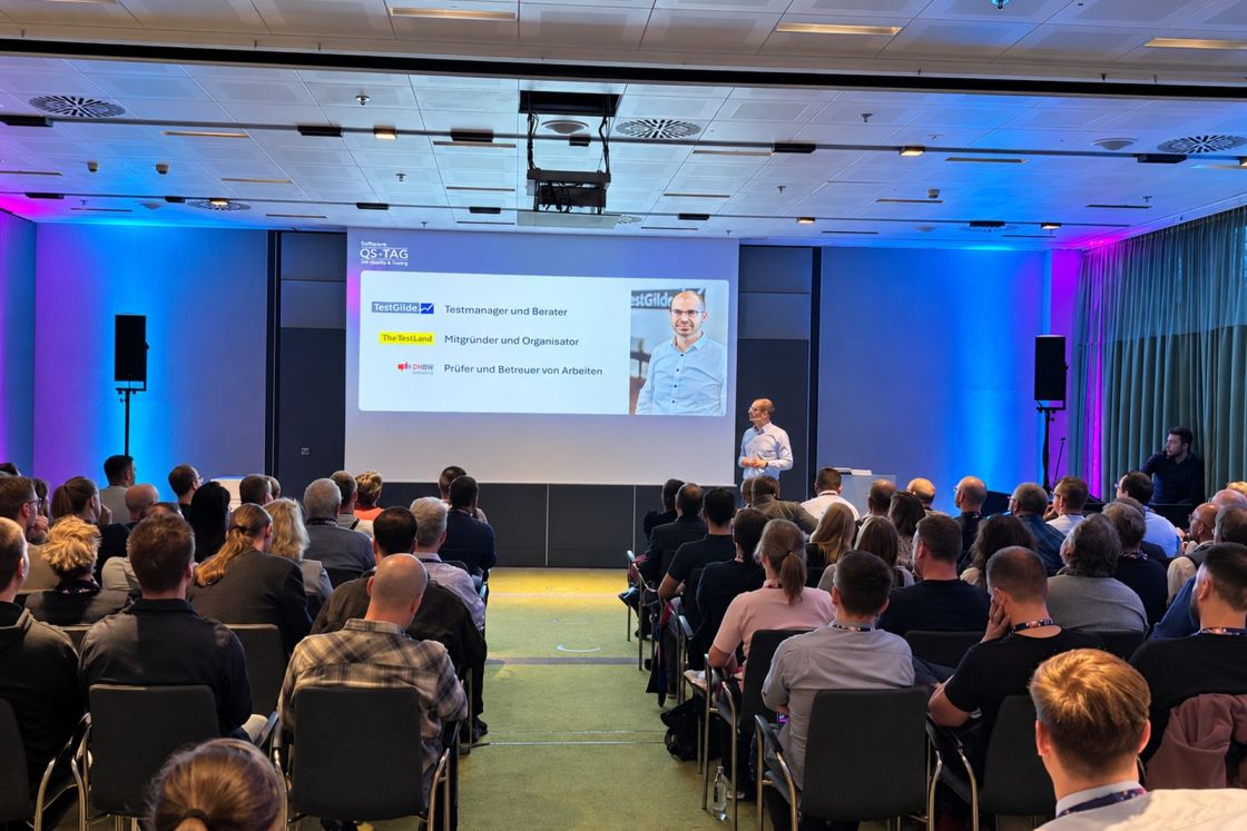 Matthias Groß spricht vor vollem Saal beim Software-QS-Tag, im Hintergrund die Folie mit seinen Rollen bei TestGilde, TestLänd und der DHBW.