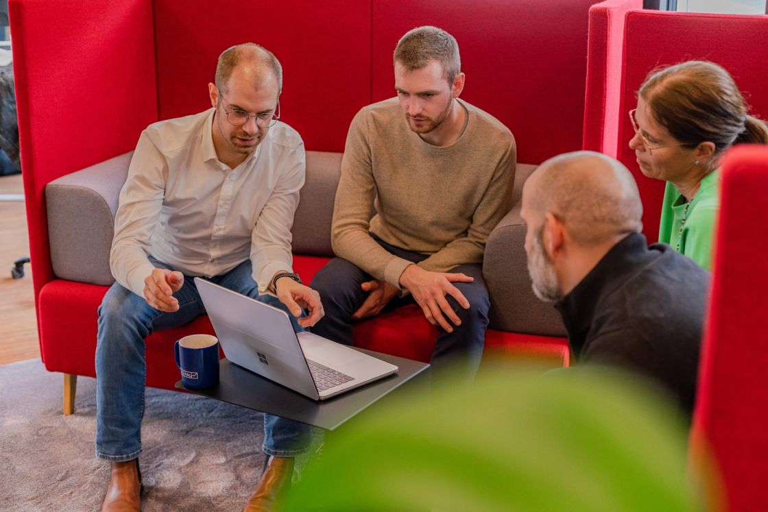 Matthias Groß bespricht im Büro der TestGilde mit einer Kollegin und 2 Kollegen etwas am Laptop, im Hintergrund rote Sitzmöbel.