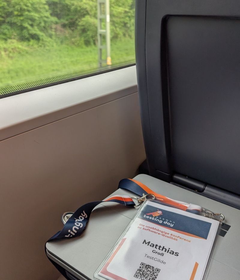 Konferenzausweis des German Testing Day mit dem Namen Matthias Groß und dem Lanyard auf dem Klapptisch im Zug, dahinter das Fenster.