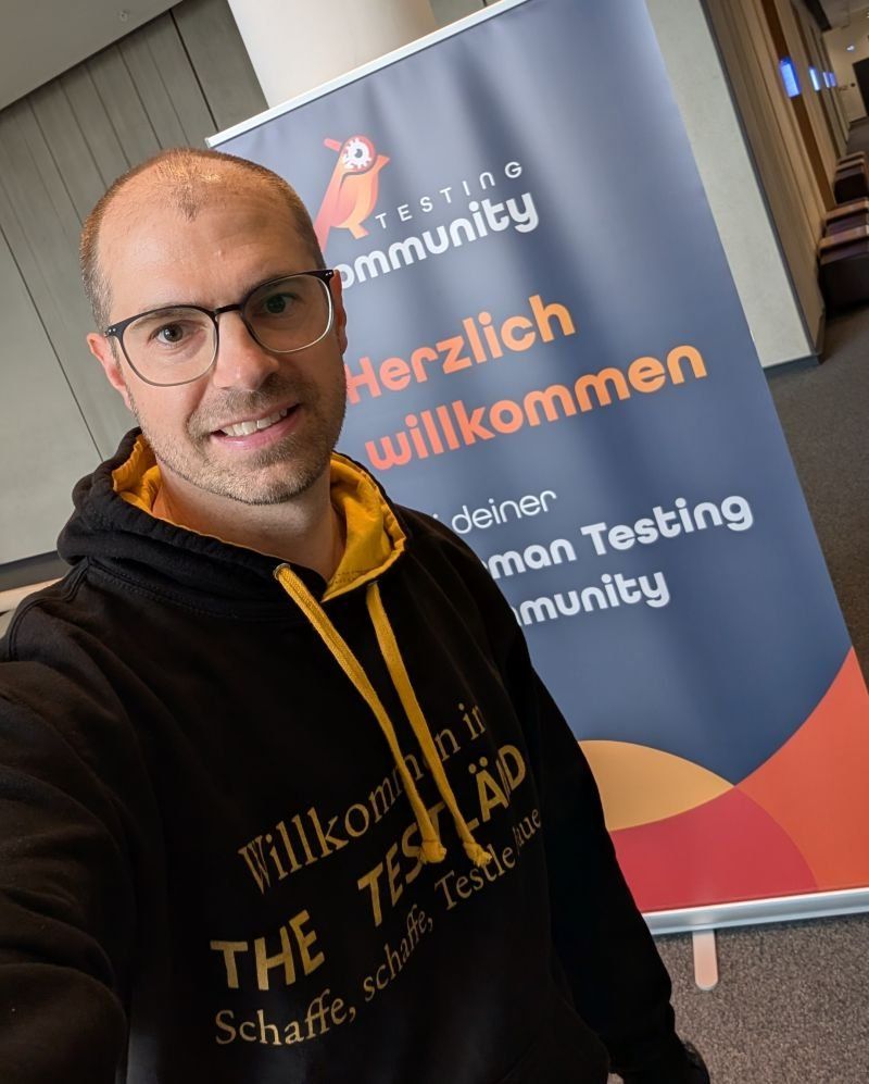 Selfie von Matthias Groß im schwarzen TestLänd-Kapuzenpulli vor dem Roll-up der German Testing Community mit der Aufschrift Herzlich willkommen.