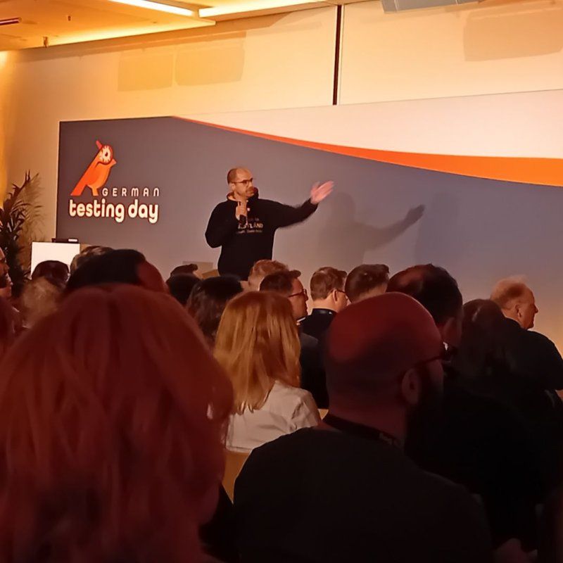 Matthias Groß im TestLänd-Kapuzenpulli spricht mit Mikrofon auf der Bühne des German Testing Day, im Vordergrund das Publikum.
