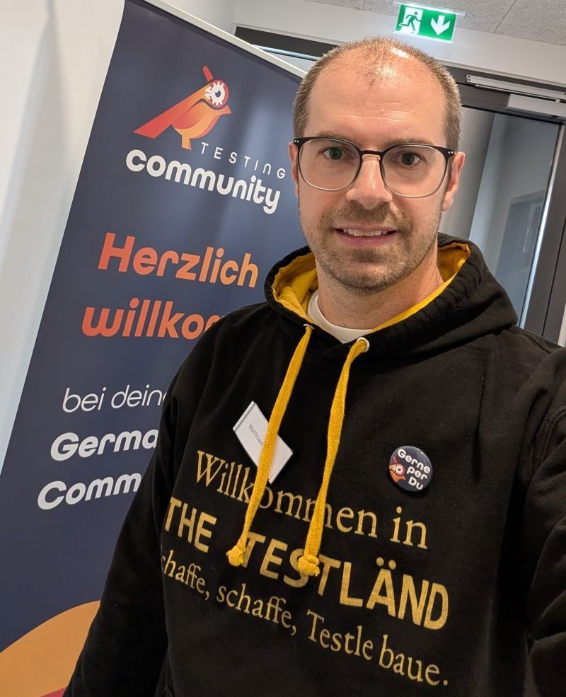 Selfie von Matthias Groß im schwarzen TestLänd-Kapuzenpulli mit dem Button Gerne per Du vor dem Roll-up der German Testing Community mit der Aufschrift Herzlich willkommen.