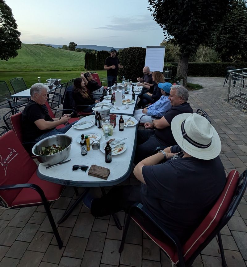 Teilnehmende des Retreats an einem langen Tisch auf einer Terrasse in der Abenddämmerung, mit Salatschüssel und Getränken. Hinten steht eine Person vor einem Flipchart, dahinter Wiesen und Hügel.