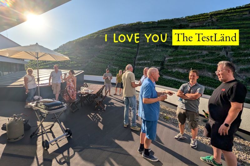 Gäste stehen bei Sonnenschein auf der Dachterrasse der TestGilde vor einem steilen Weinberg, darüber der Schriftzug I love you und der Name TestLänd.