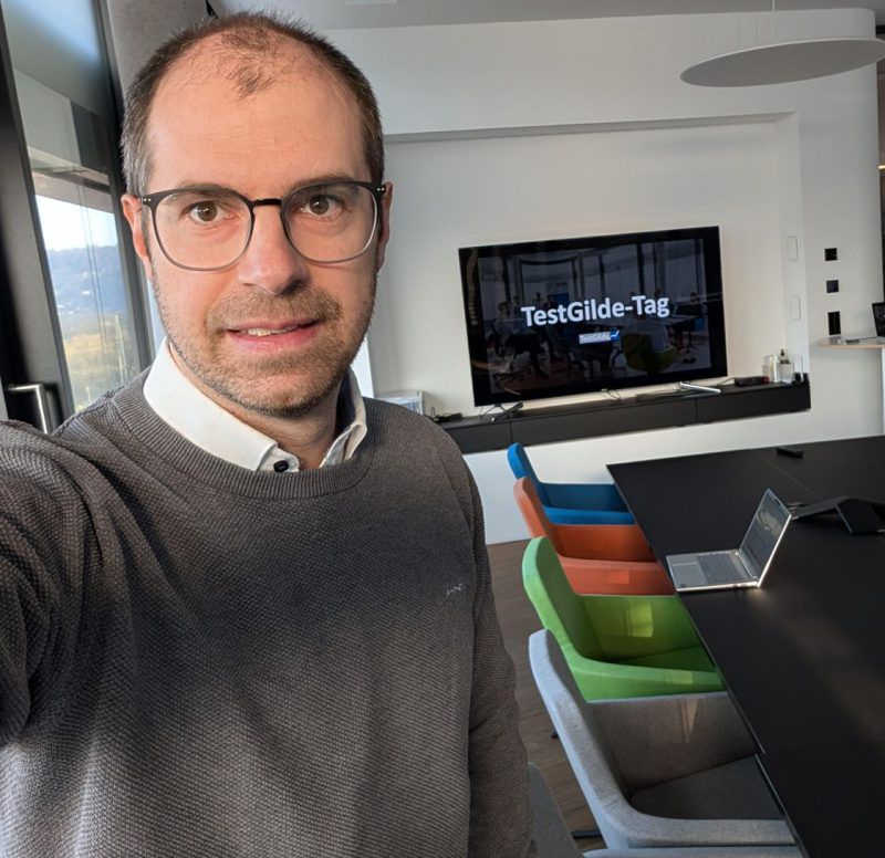 Selfie von Matthias Groß im TestGilde-Büro. Hinter ihm zeigt ein Bildschirm den Schriftzug TestGilde-Tag, rechts stehen bunte Stühle an einem langen Tisch mit aufgeklapptem Laptop.