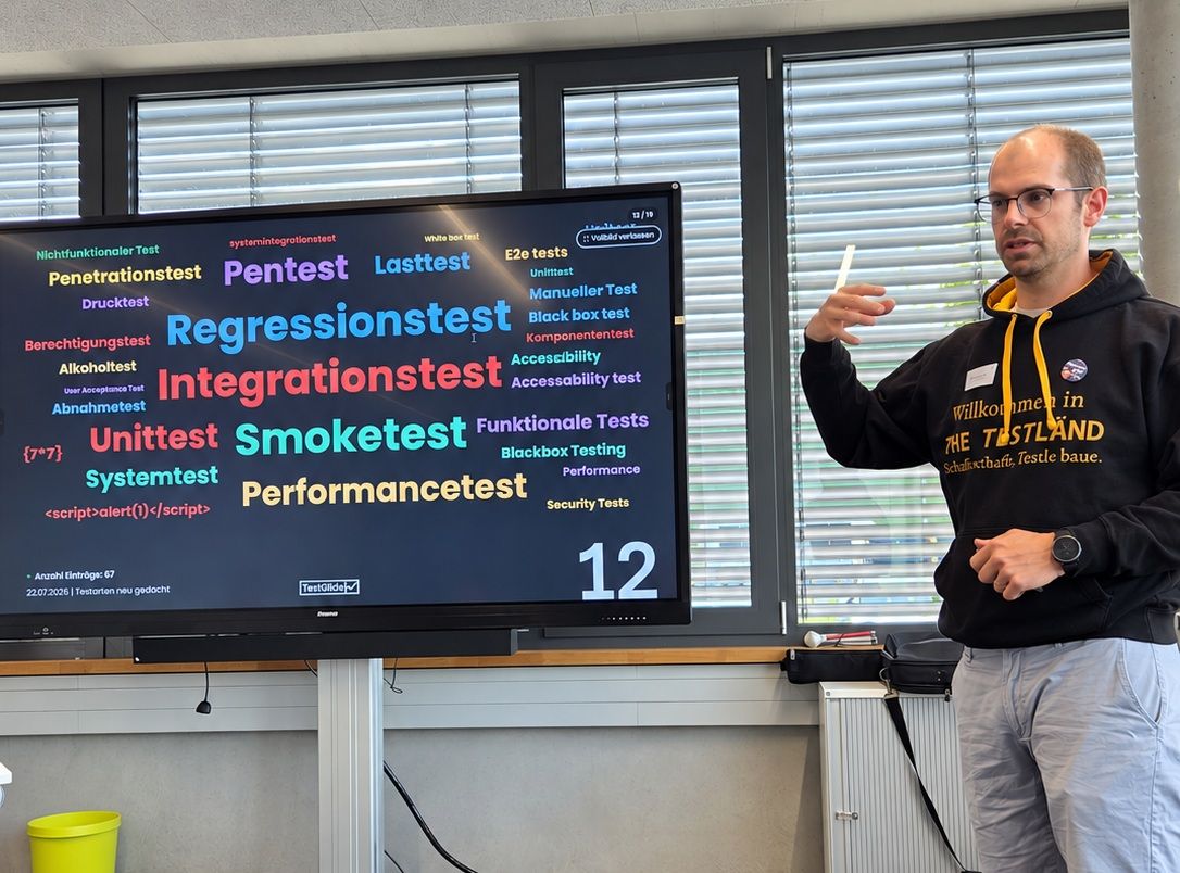Matthias Groß hält den Vortrag Testarten neu gedacht vor einem Bildschirm mit einer Live-Wortwolke aus Begriffen wie Regressionstest, Integrationstest, Unittest, Smoketest und Performancetest.