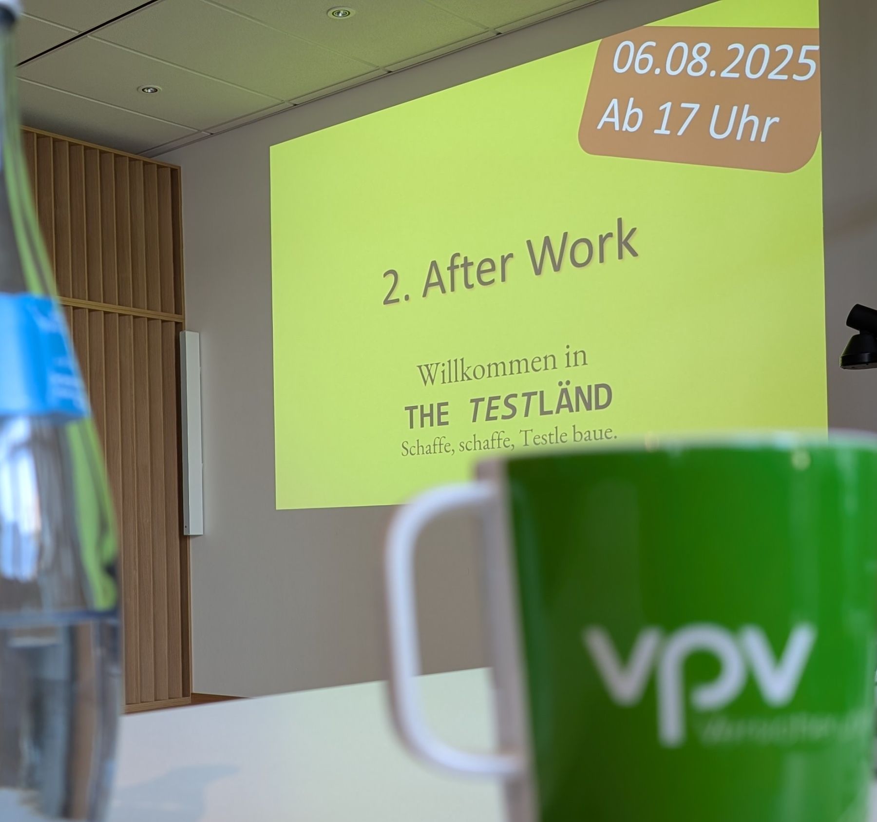 Projektion mit der Folie „2. After Work, Willkommen in The TestLänd, 06.08.2025, ab 17 Uhr“, im Vordergrund unscharf eine grüne Tasse der VPV.