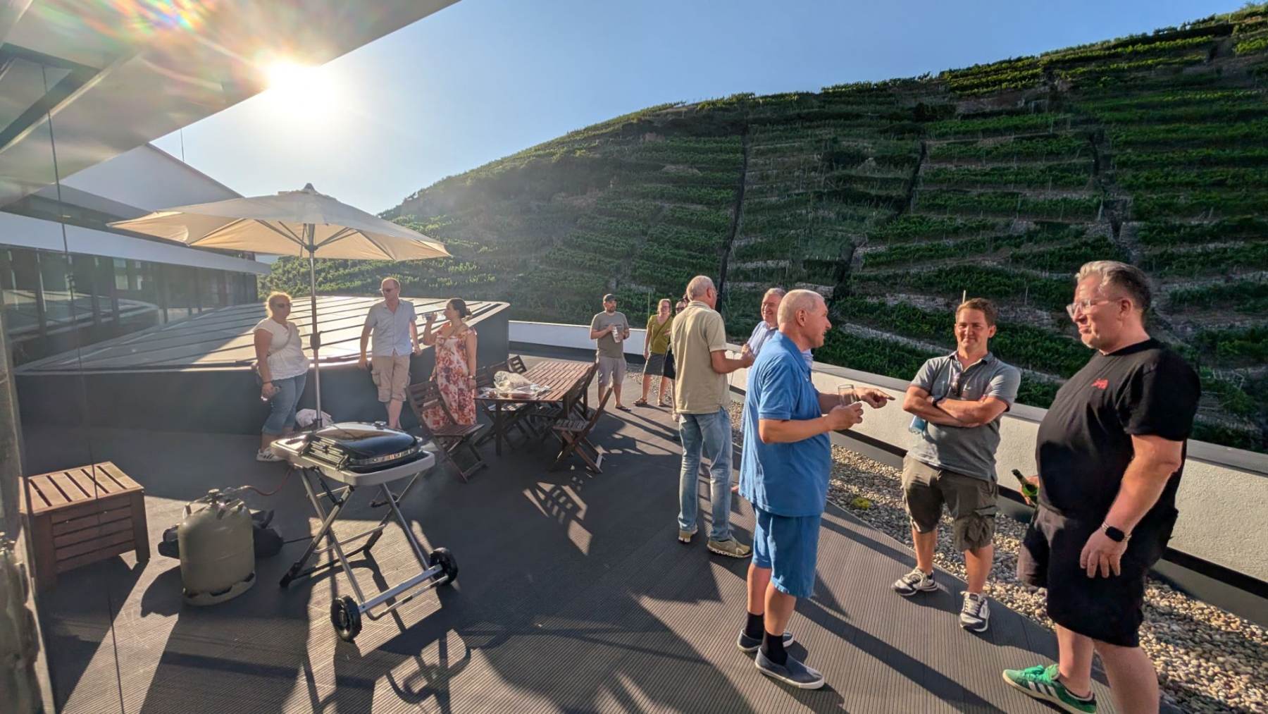 Gäste stehen bei Sonnenschein mit Getränken auf der Dachterrasse der TestGilde, im Hintergrund ein steiler Weinberg.