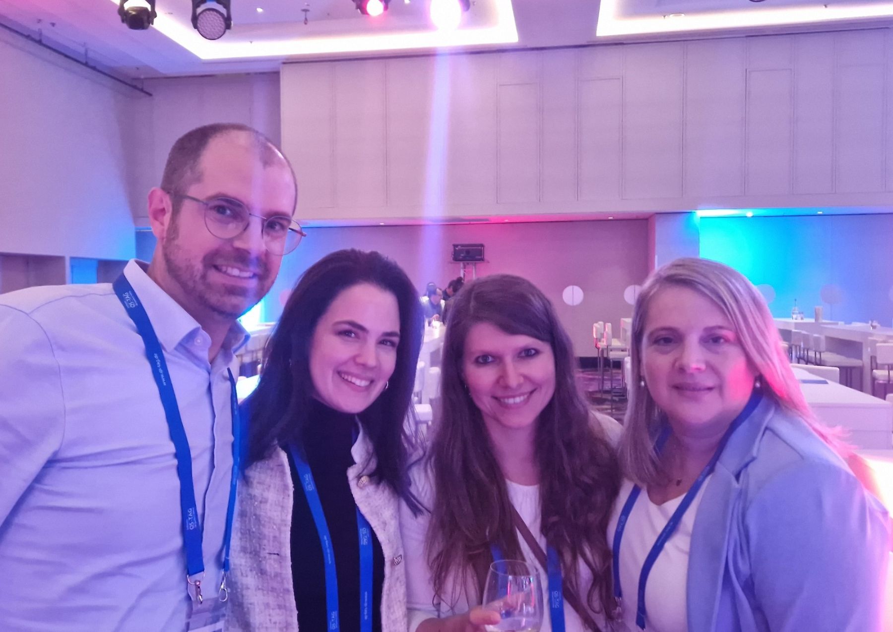 Matthias Groß, Laura de Michele, Julia Leitner und Anita Hellwig mit Umhängebändern des Software-QS-Tag lächeln in die Kamera, im Hintergrund ein Saal in blauem und rosa Licht.