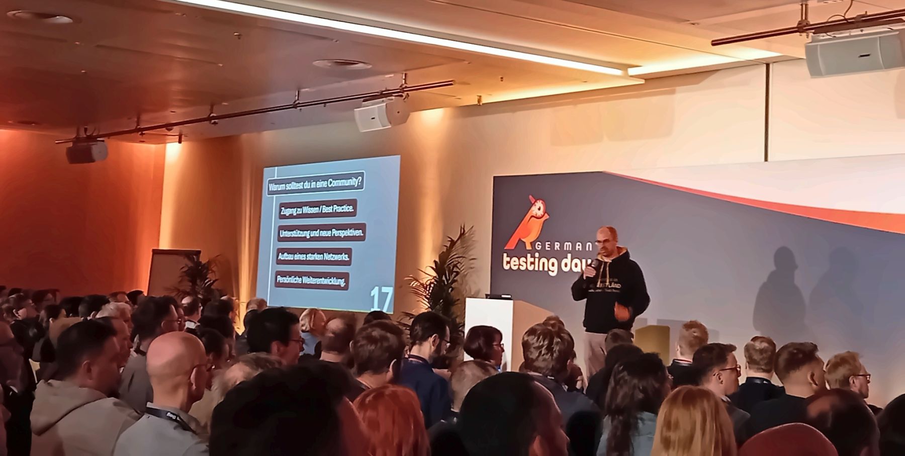 Matthias Groß hält den Impulsvortrag The TestLänd vor vollem Saal beim German Testing Day, im Hintergrund die Folie mit den Gründen für die Mitgliedschaft in einer Community.